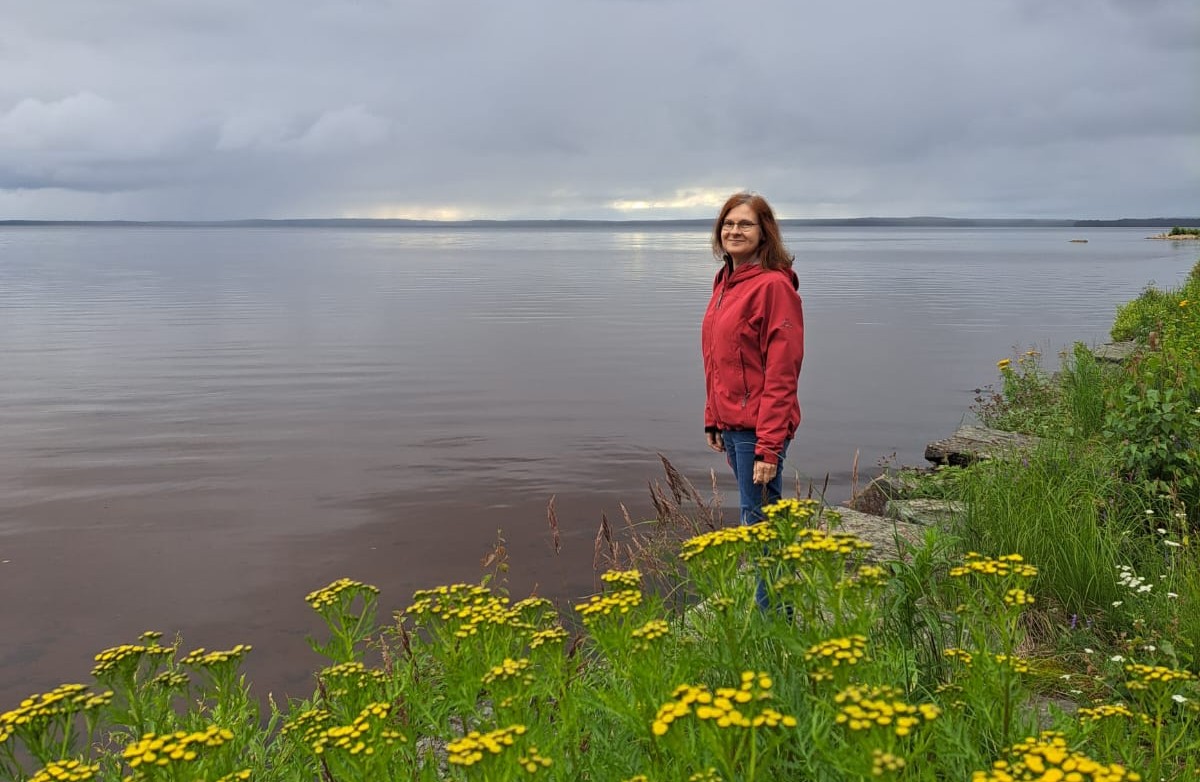 Blogin kirjoittaja seisomassa Oulujärven rannassa, etualalla keltaisia kukkia.